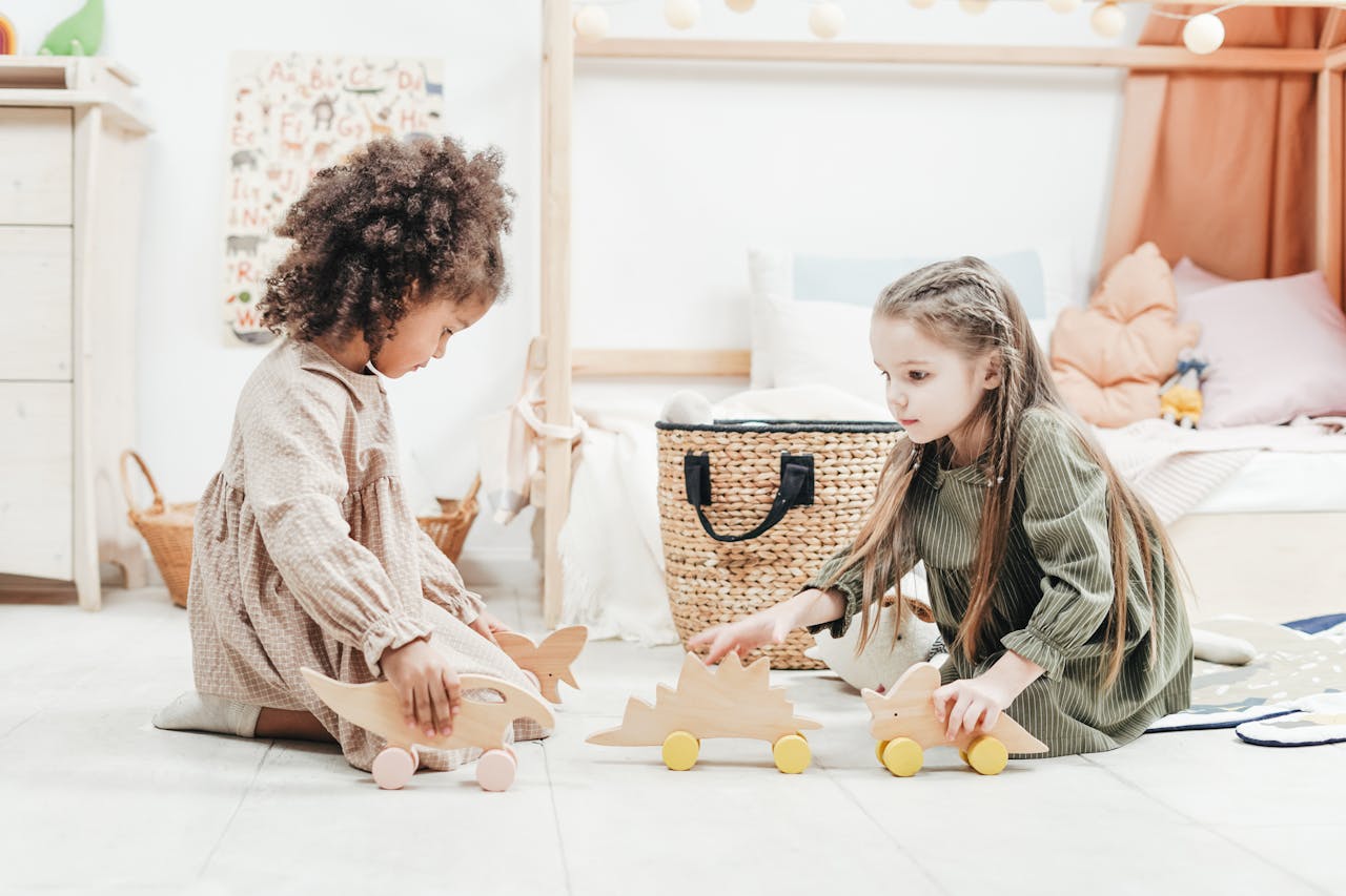 Two young girls playing with wooden dinosaur toys in an indoor playroom setting.
